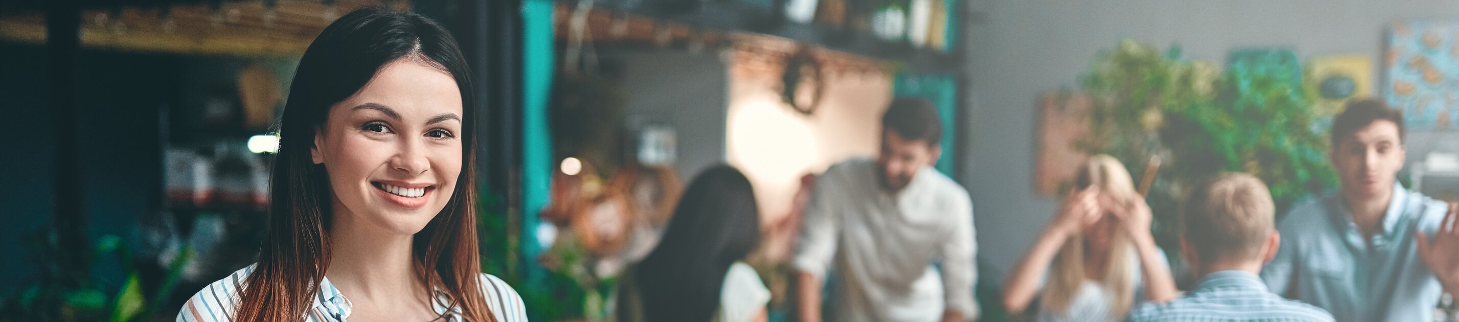 Junge Frau mit Laptop lächelt im kreativen Büro während Teammeeting im Hintergrund