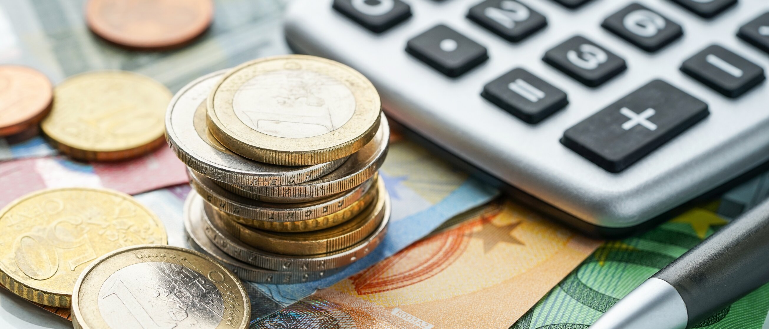 Gestapelte Euromünzen und Geldscheine neben Taschenrechner und Stift als Symbol für Kalkulation und Finanzplanung