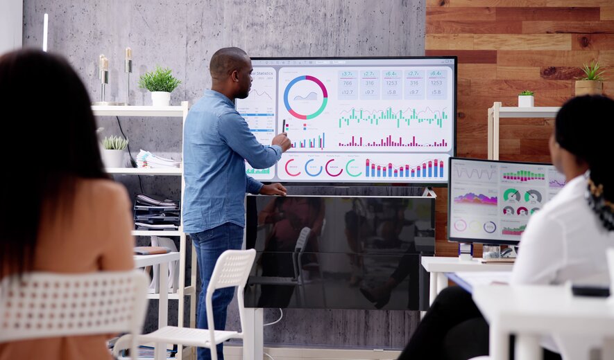 Teammeeting mit Präsentation, bei der ein Mitarbeiter Diagramme und Daten auf einem großen Bildschirm erklärt