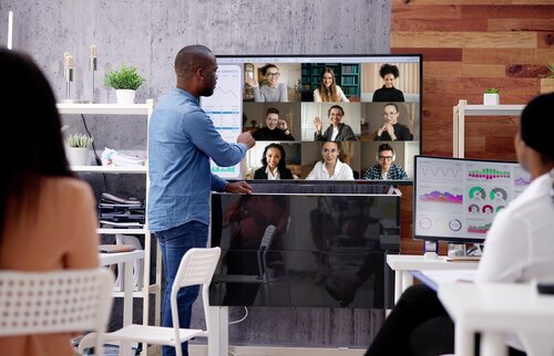 Trainer steht vor einem Bildschirm mit Teilnehmenden im Videocall und spricht zu einer kleinen Gruppe im Raum – hybrider Unterricht mit Online- und Präsenzteilnehmenden.