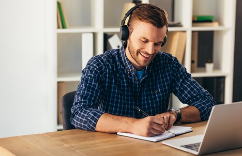 Mann lernt online am Laptop mit Kopfhörern und Notizbuch.