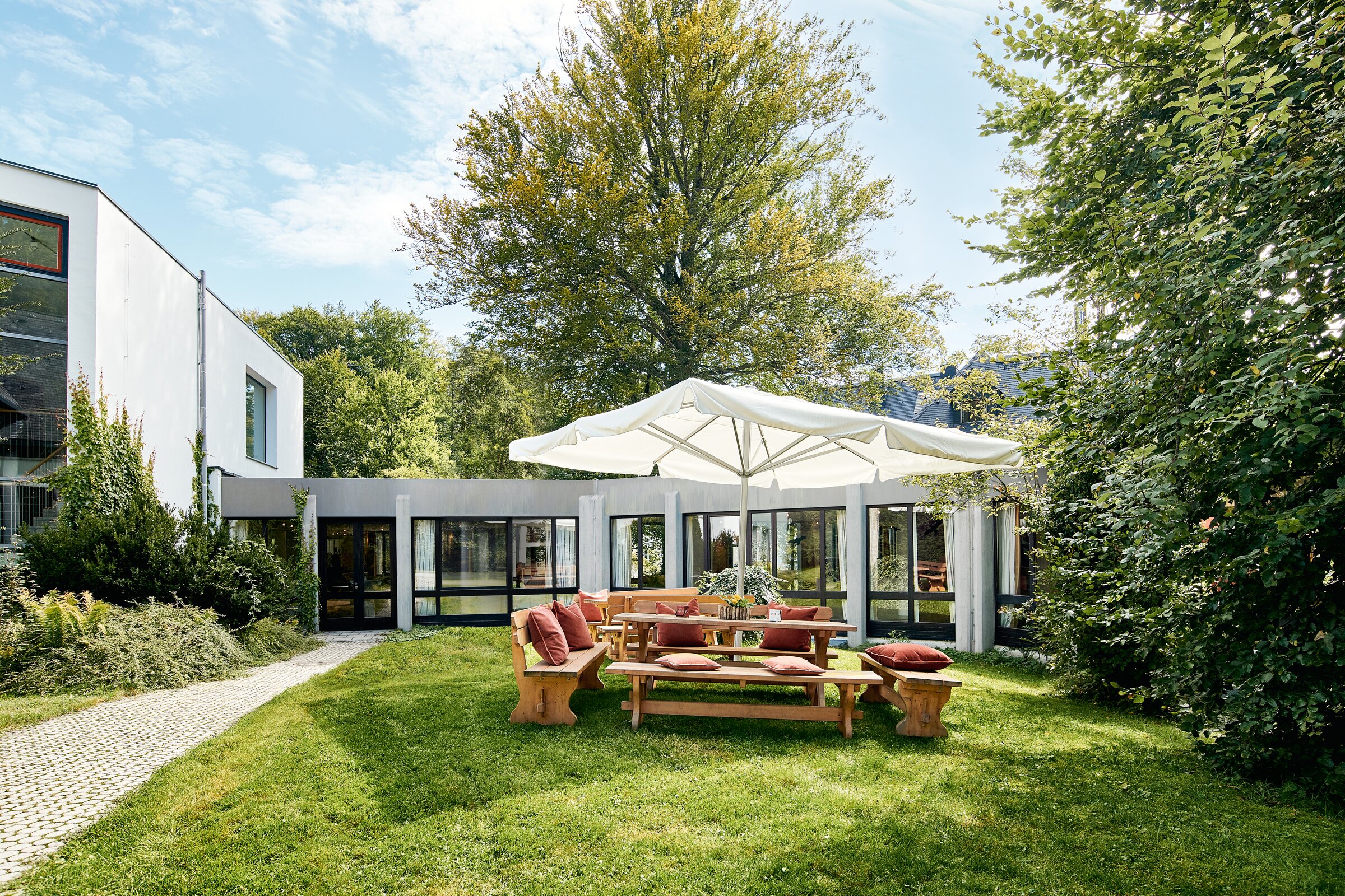 Sonnige Gartenterrasse der Akademie Westerham mit Sitzgruppen und Sonnenschirm, umgeben von Bäumen und Wiese