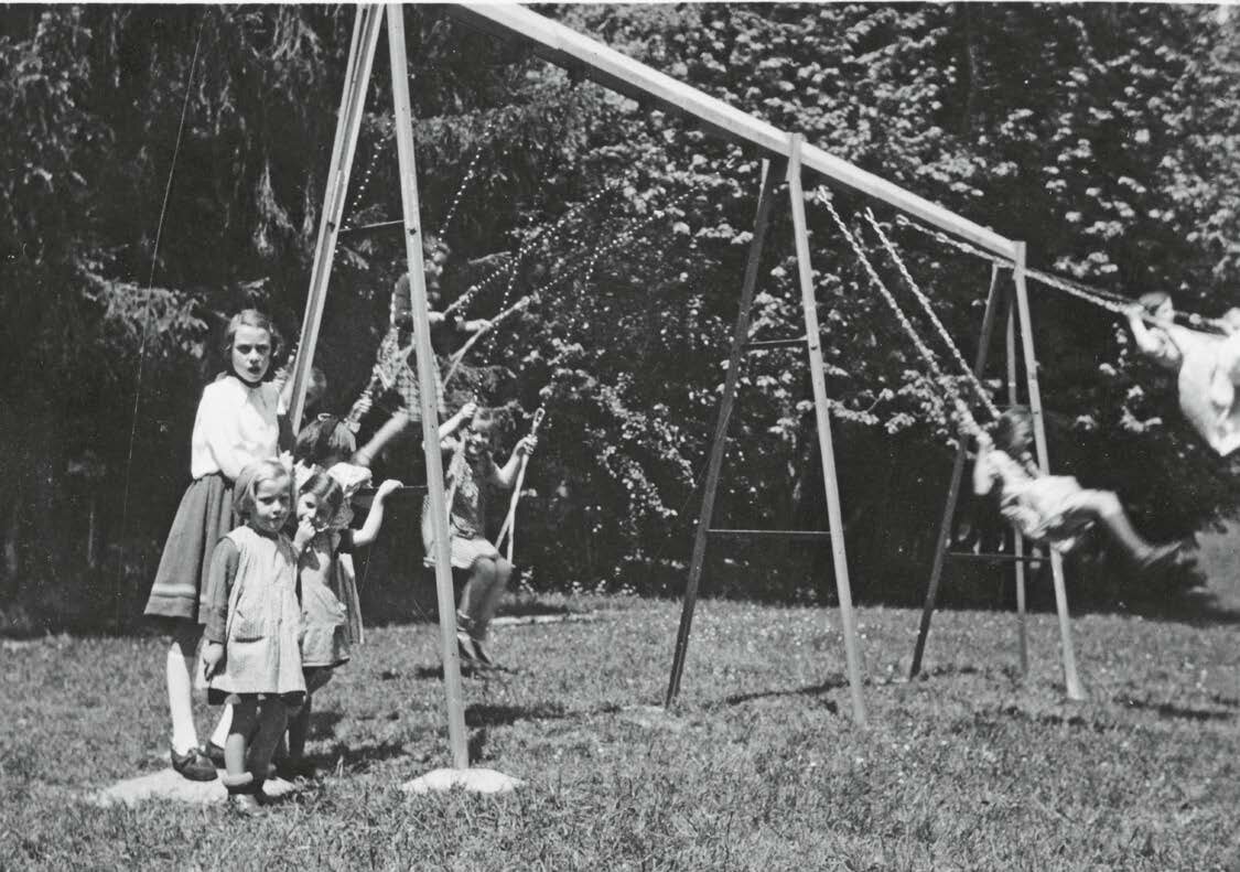 Schwarz-Weiß-Fotografie spielender Kinder auf einer Schaukel im Garten des Kinderkurheims Westerham, umgeben von Bäumen