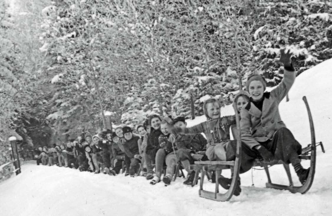 Historische Schwarz-Weiß-Fotografie von Kindern beim Rodeln auf einer verschneiten Waldstraße