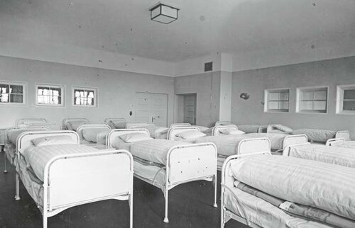 Schwarz-Weiß-Aufnahme eines hellen Schlafsaals im Kinderkurheim Westerham der 1920er Jahre mit zahlreichen ordentlich aufgereihten Metallbetten und großen Fenstern