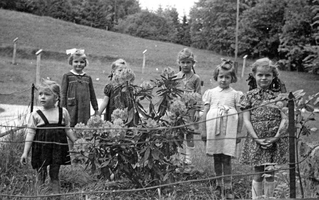 Schwarz-Weiß-Fotografie von sechs Mädchen im Garten des Kinderkurheims Westerham, stehend vor einem Blumenbeet mit Hügelwiese und Bäumen im Hintergrund