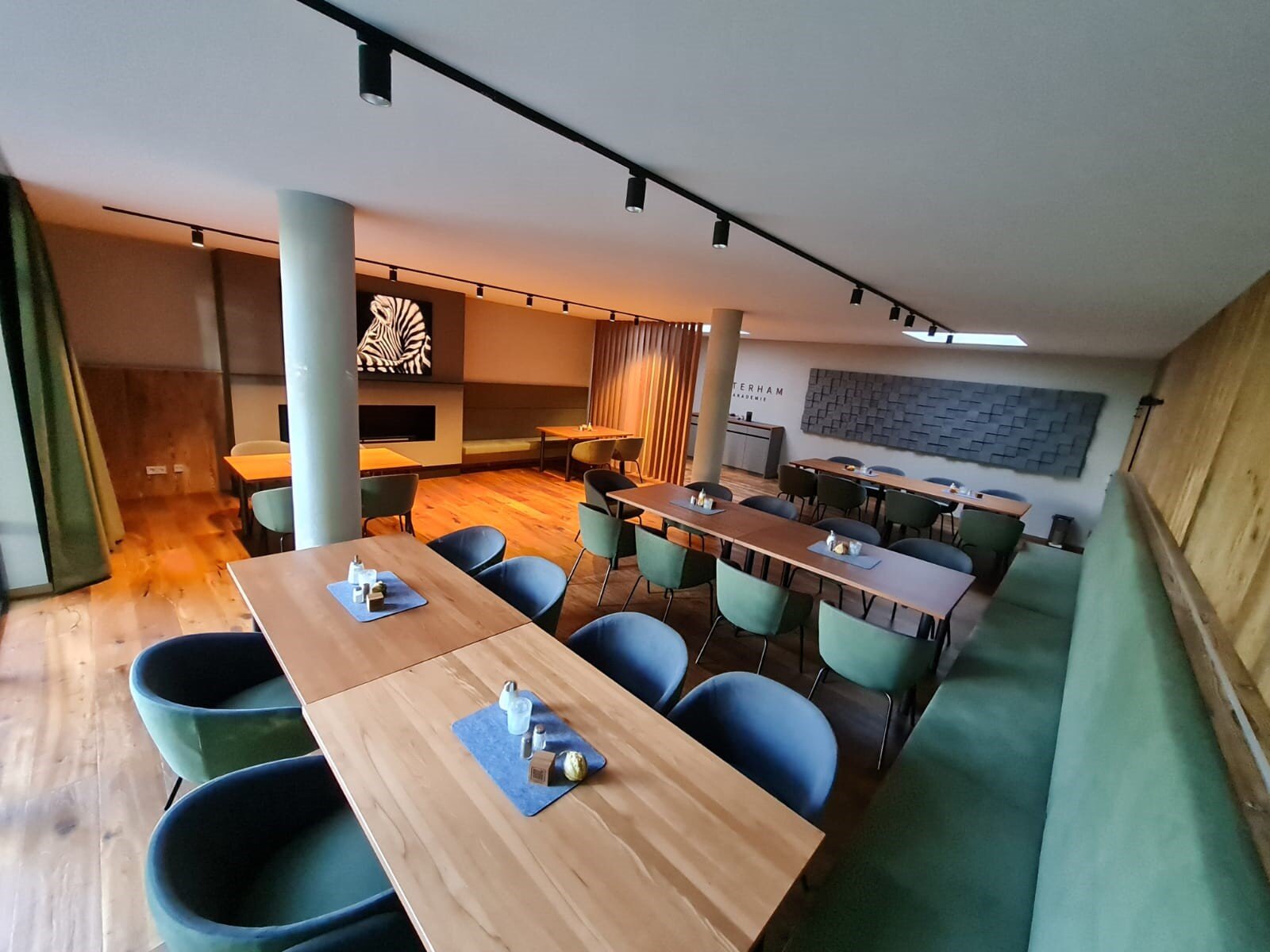 Heller, modern eingerichteter Seminar- und Gastraum mit Holztischen, gepolsterten Stühlen, Akustikpaneelen an der Wand und Kaminbereich im Kaminstüberl Westerham.