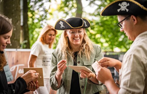 Mehrere Personen mit Piratenhüten stehen im Freien, lachen und lösen gemeinsam Rätsel auf Karten