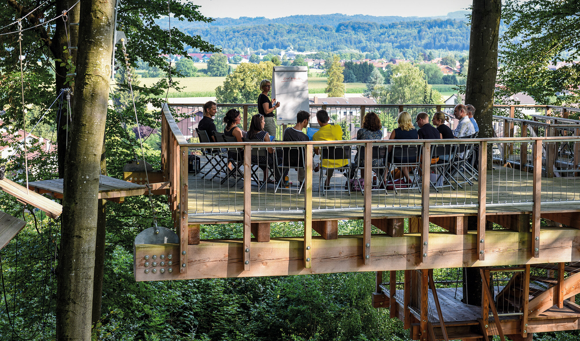 Teilnehmergruppe auf der Teamplattform der IHK Akademie Westerham inmitten hoher Bäume mit Blick ins Tal