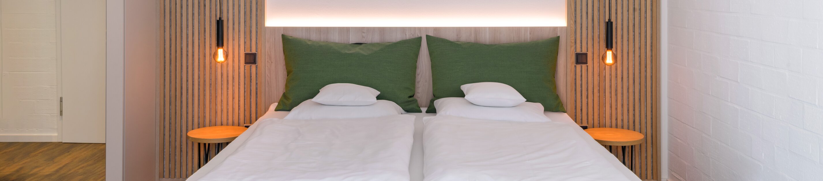 Ein modernes, schlichtes Doppelzimmer mit grünem Bett, warmen Holzakzenten und stimmungsvoller Beleuchtung.