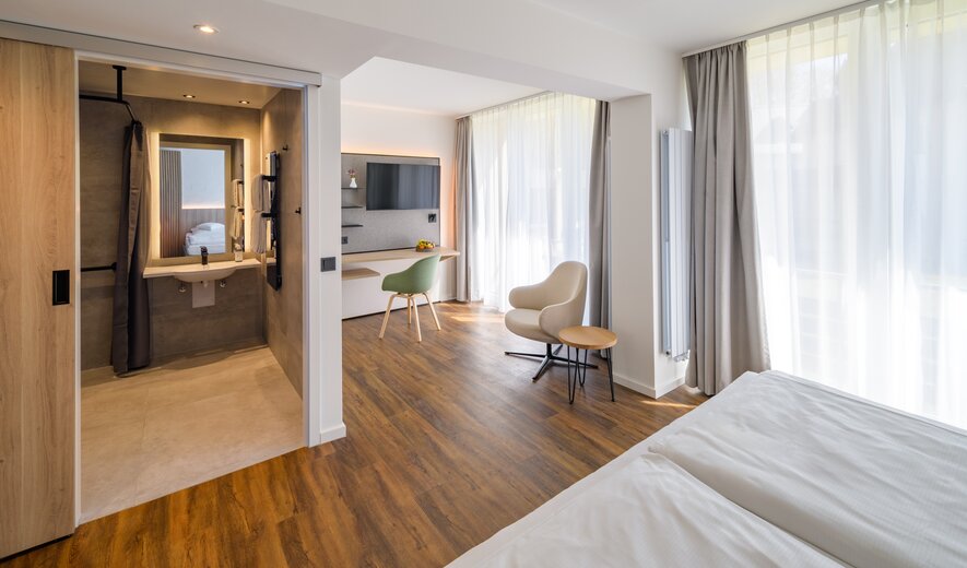 Ein helles, modernes Zimmer mit viel Tageslicht, warmem Holzboden, offenem Badbereich und stilvollen Sitz- und Arbeitsplätzen.