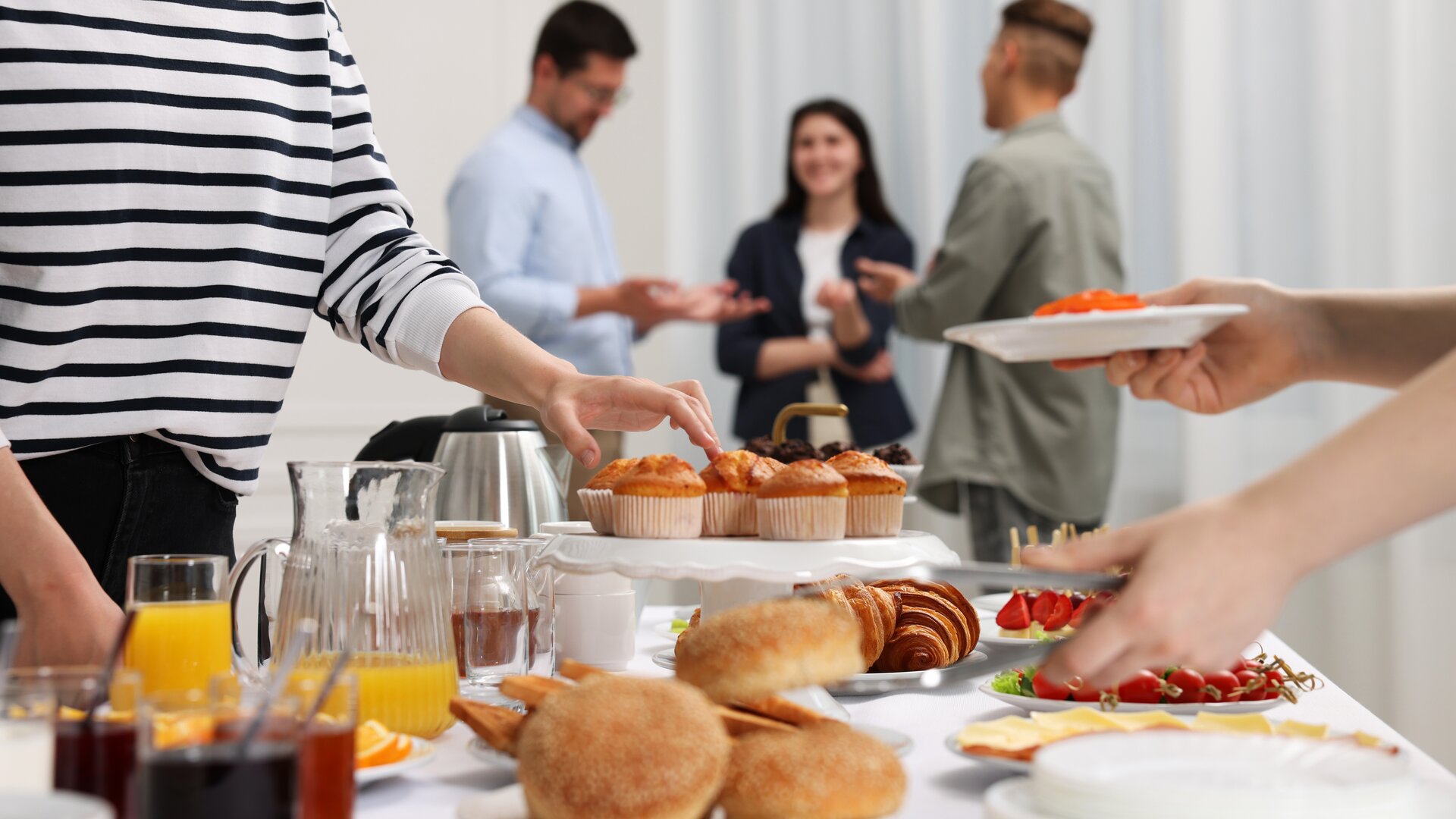 Ein reichhaltiges Buffet mit Gebäck, Aufschnitt und Säften, während sich im Hintergrund mehrere Personen ungezwungen unterhalten.
