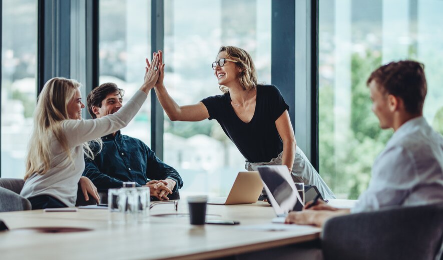 Zwei Personen geben sich lachend ein High-Five, während ihre Kolleginnen und Kollegen am Konferenztisch erfreut zusehen.