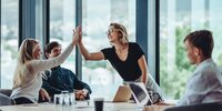 Zwei Personen geben sich lachend ein High-Five, während ihre Kolleginnen und Kollegen am Konferenztisch erfreut zusehen.