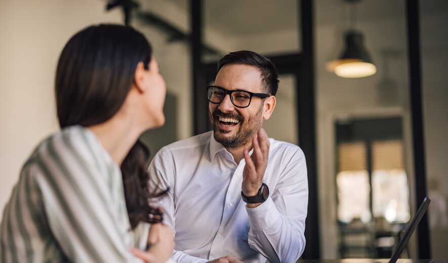 Ein Mann und eine Frau sitzen sich in einem Büro gegenüber, unterhalten sich angeregt und lachen gemeinsam.