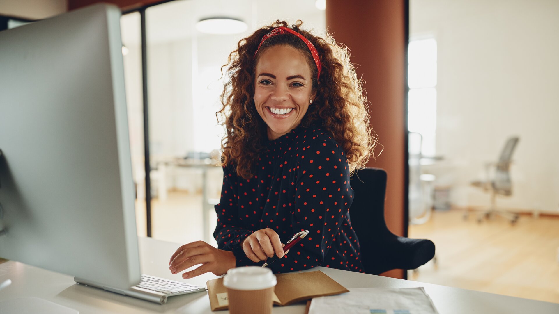 Auf dem Bild sieht man eine fröhliche Frau an einem Schreibtisch im Büro, die lächelnd in die Kamera schaut und gerade an ihrem Computerarbeitsplatz sitzt. 