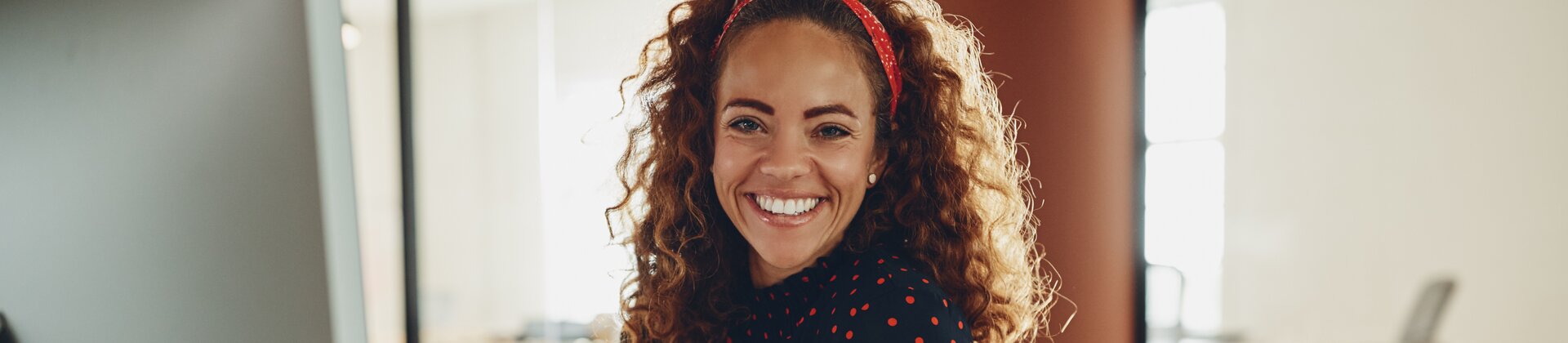 Auf dem Bild sieht man eine fröhliche Frau an einem Schreibtisch im Büro, die lächelnd in die Kamera schaut und gerade an ihrem Computerarbeitsplatz sitzt. 