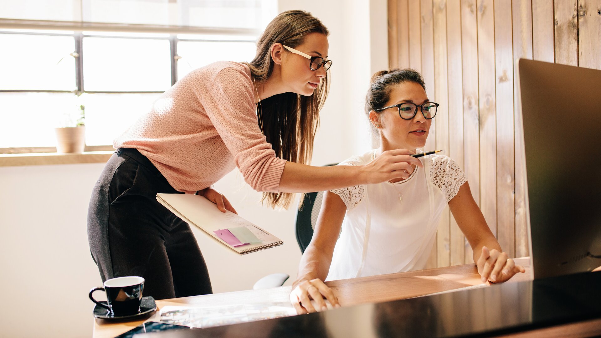 Zwei Frauen arbeiten gemeinsam an einem Computer, wobei eine der beiden der anderen etwas auf dem Bildschirm zeigt.