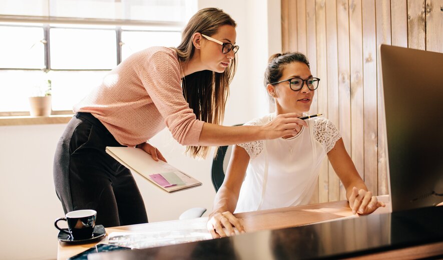 Zwei Frauen arbeiten gemeinsam an einem Computer, wobei eine der beiden der anderen etwas auf dem Bildschirm zeigt.