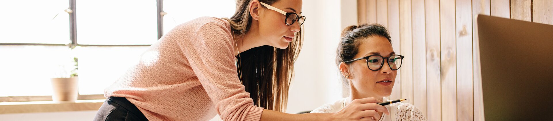 Zwei Frauen arbeiten gemeinsam an einem Computer, wobei eine der beiden der anderen etwas auf dem Bildschirm zeigt.