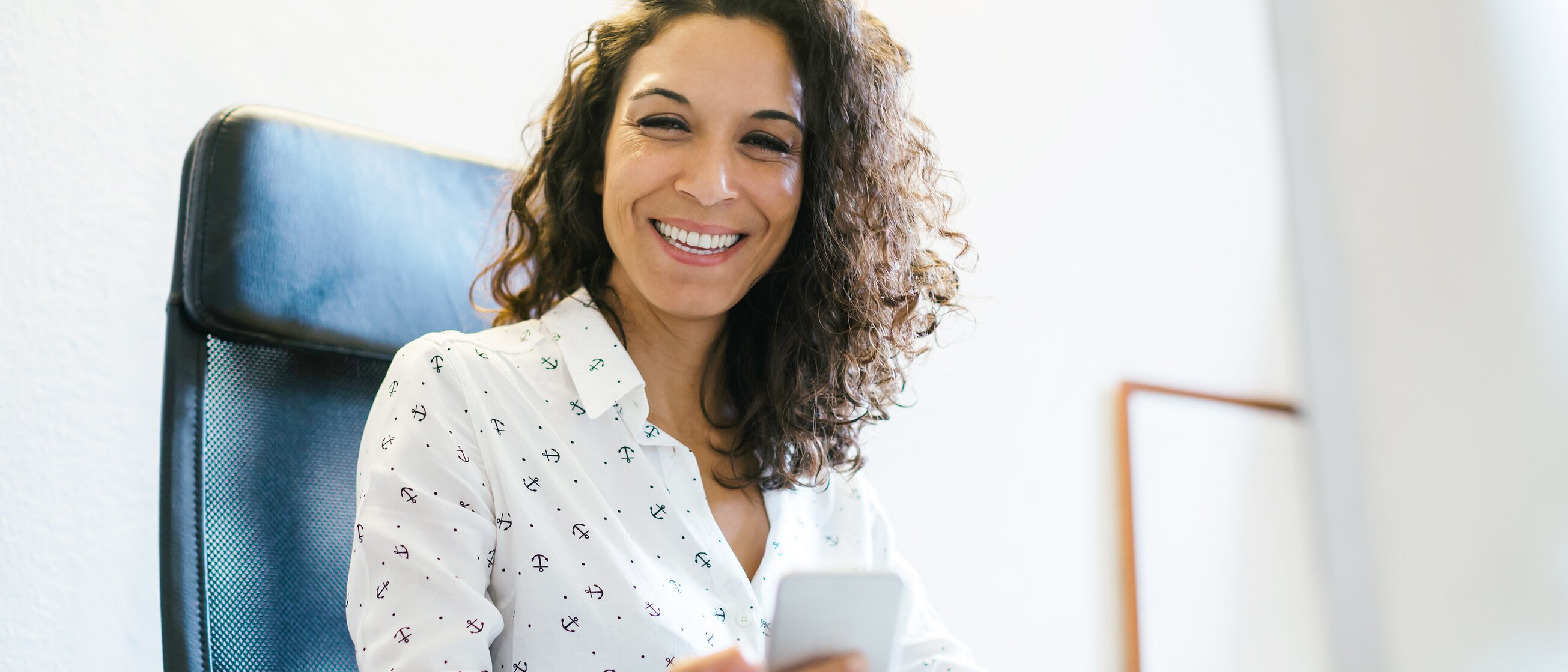 Frau sitzt lächelnd im Büro und hält ein Smartphone in der Hand – Symbol für modernes Office Management und digitale Arbeitsorganisation.