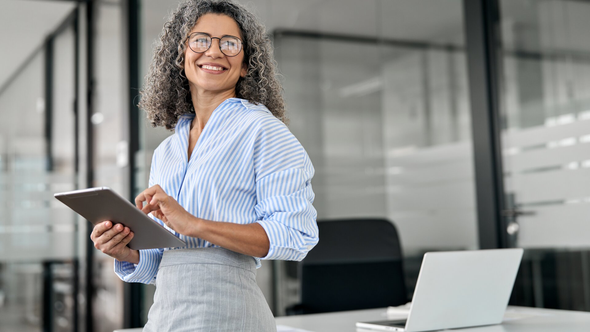 Eine freundlich wirkende Frau steht in einem modernen Büro, hält ein Tablet in den Händen und lächelt selbstbewusst in Richtung Kamera.