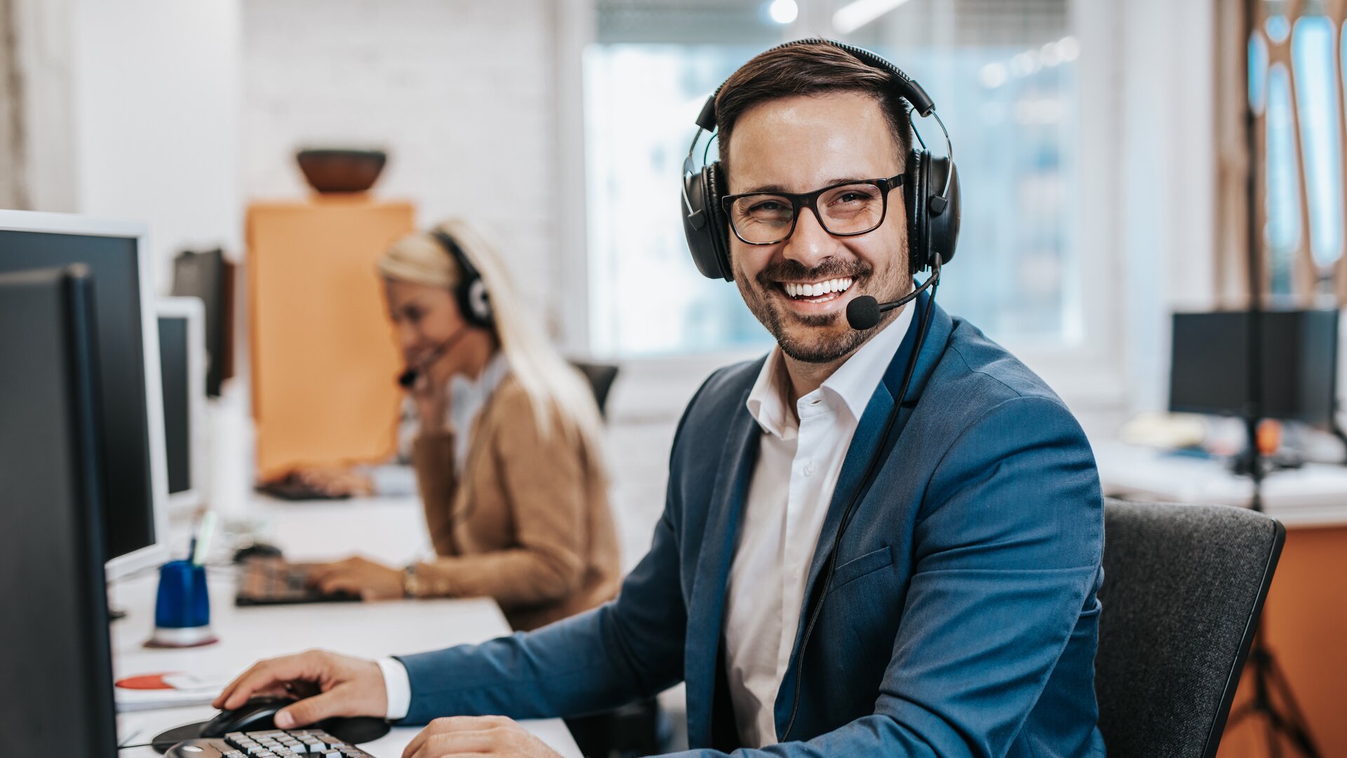Ein lächelnder Mann mit Headset sitzt an einem Computerarbeitsplatz in einem Großraumbüro und wirkt, als führe er ein Service- oder Supportgespräch.