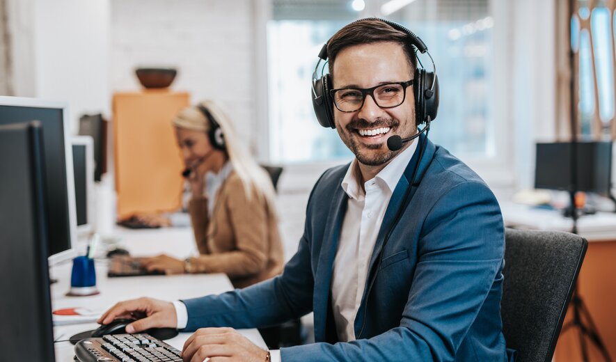 Ein lächelnder Mann mit Headset sitzt an einem Computerarbeitsplatz in einem Großraumbüro und wirkt, als führe er ein Service- oder Supportgespräch.