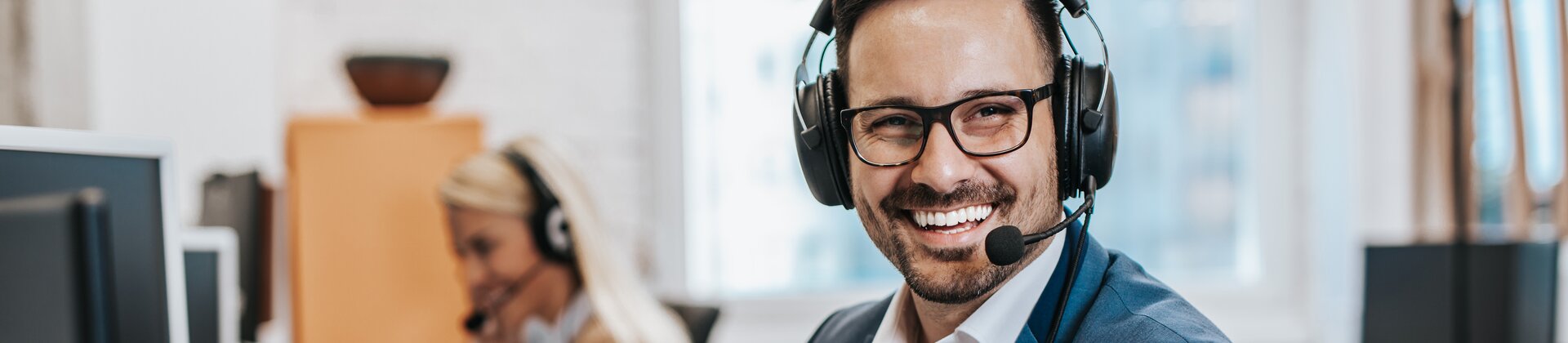 Ein lächelnder Mann mit Headset sitzt an einem Computerarbeitsplatz in einem Großraumbüro und wirkt, als führe er ein Service- oder Supportgespräch.