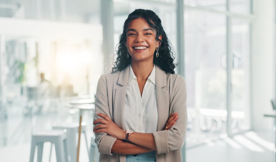 Auf dem Bild steht eine lächelnde Frau mit verschränkten Armen in einem modernen, hellen Büro und wirkt selbstbewusst und professionell.
