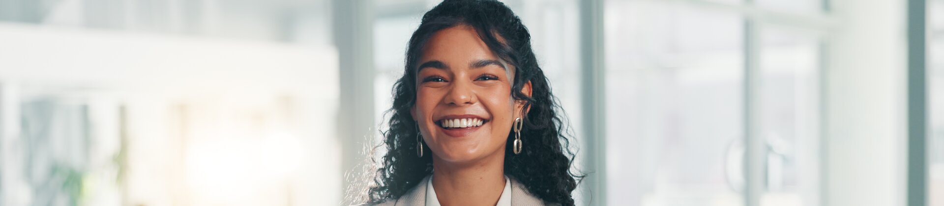 Auf dem Bild steht eine lächelnde Frau mit verschränkten Armen in einem modernen, hellen Büro und wirkt selbstbewusst und professionell.
