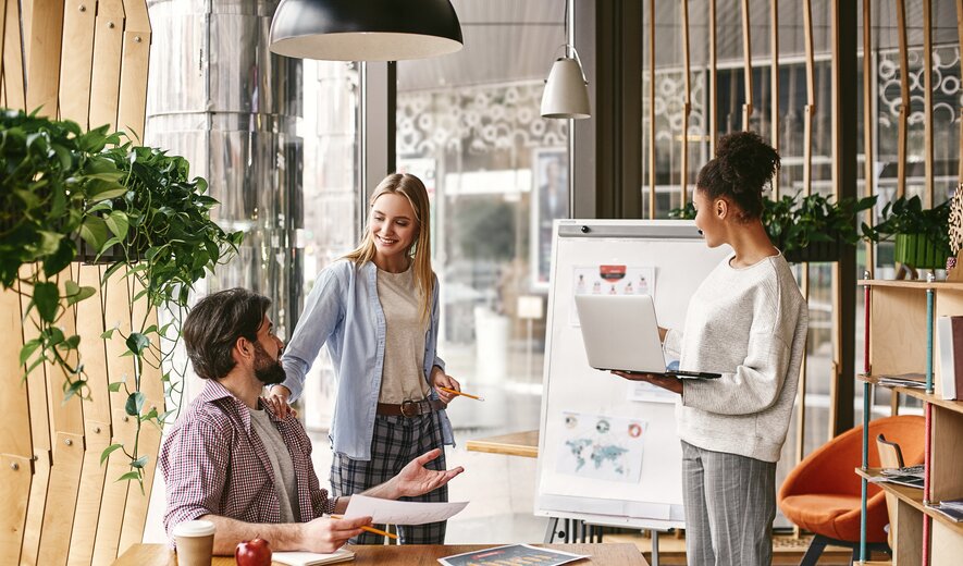 Drei Personen arbeiten in einer modernen, gemütlichen Umgebung zusammen, während eine Präsentation auf einem Flipchart gezeigt wird.