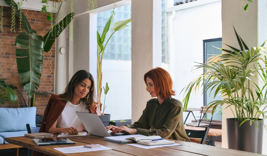 Zwei Frauen sitzen an einem Tisch in einem hellen, grünen Arbeitsumfeld und arbeiten gemeinsam an einem Laptop.