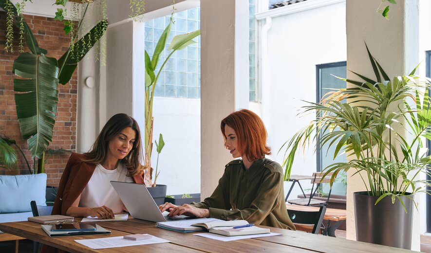 Zwei Frauen sitzen an einem Tisch in einem hellen, grünen Arbeitsumfeld und arbeiten gemeinsam an einem Laptop.