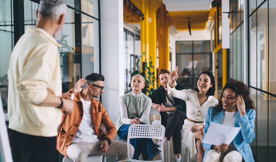 Eine kleine Gruppe von Menschen sitzt in einem modernen, farbenfrohen Seminarraum und beteiligt sich aktiv an einer Diskussion mit einer stehenden Person vorne.