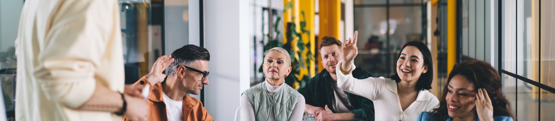 Eine kleine Gruppe von Menschen sitzt in einem modernen, farbenfrohen Seminarraum und beteiligt sich aktiv an einer Diskussion mit einer stehenden Person vorne.