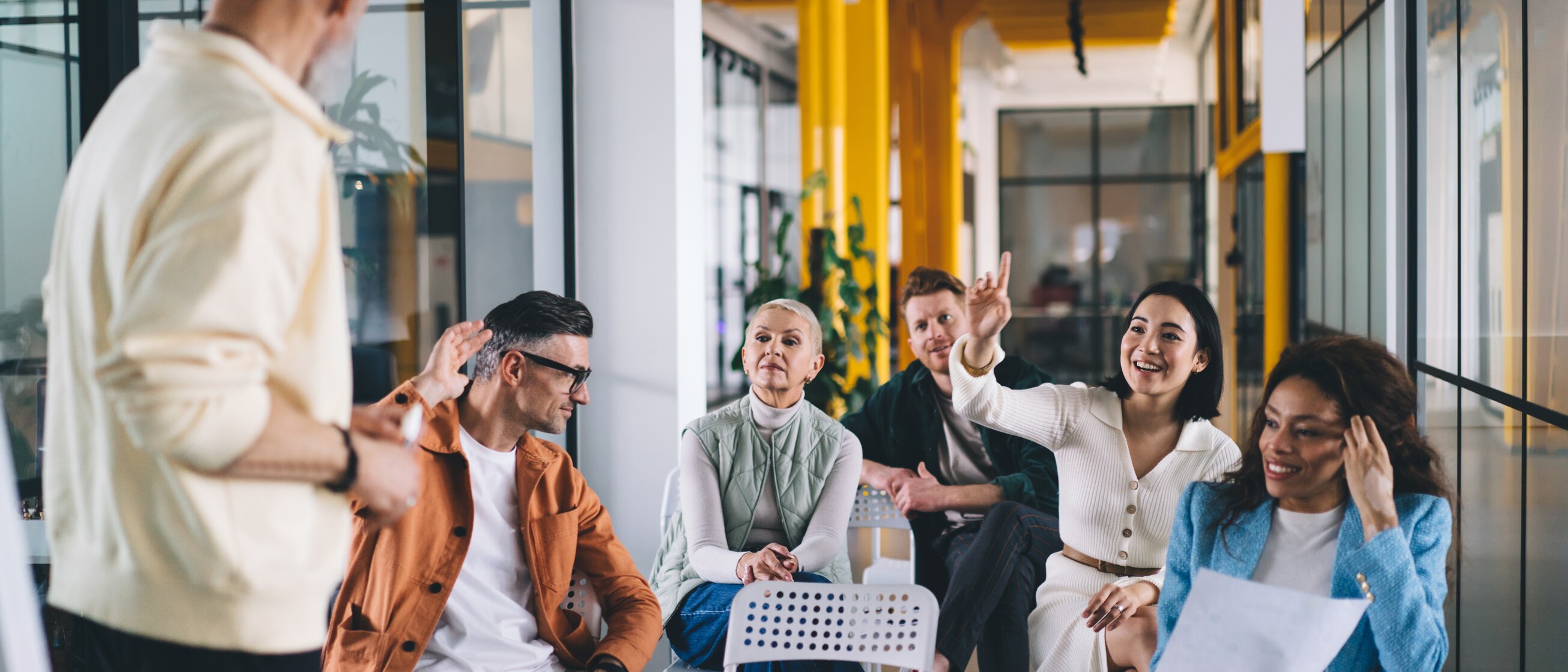 Eine kleine Gruppe von Menschen sitzt in einem modernen, farbenfrohen Seminarraum und beteiligt sich aktiv an einer Diskussion mit einer stehenden Person vorne.