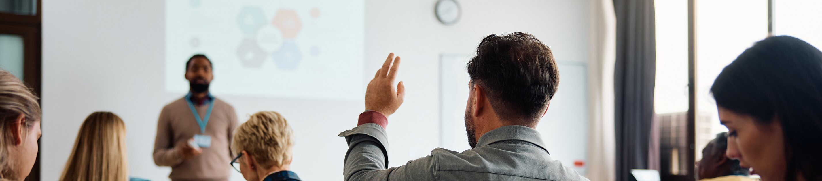 Eine Person hebt in einem Seminarraum die Hand, während der Vortragende vorne präsentiert und andere Teilnehmende mitschreiben.