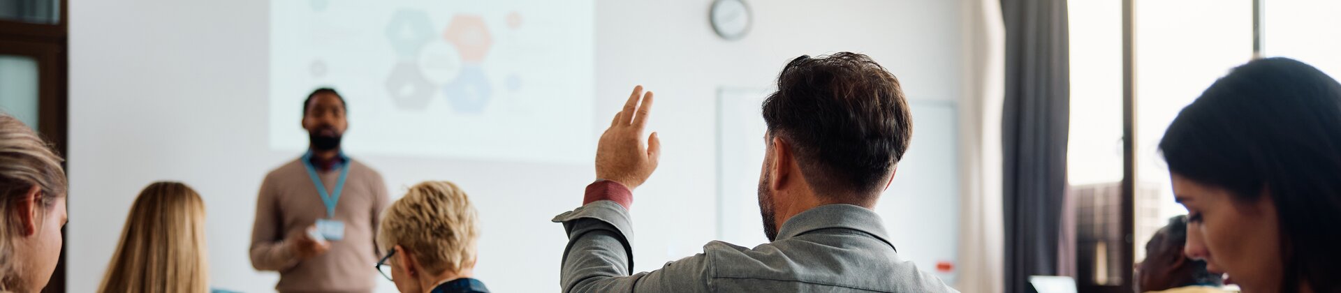 Eine Person hebt in einem Seminarraum die Hand, während der Vortragende vorne präsentiert und andere Teilnehmende mitschreiben.