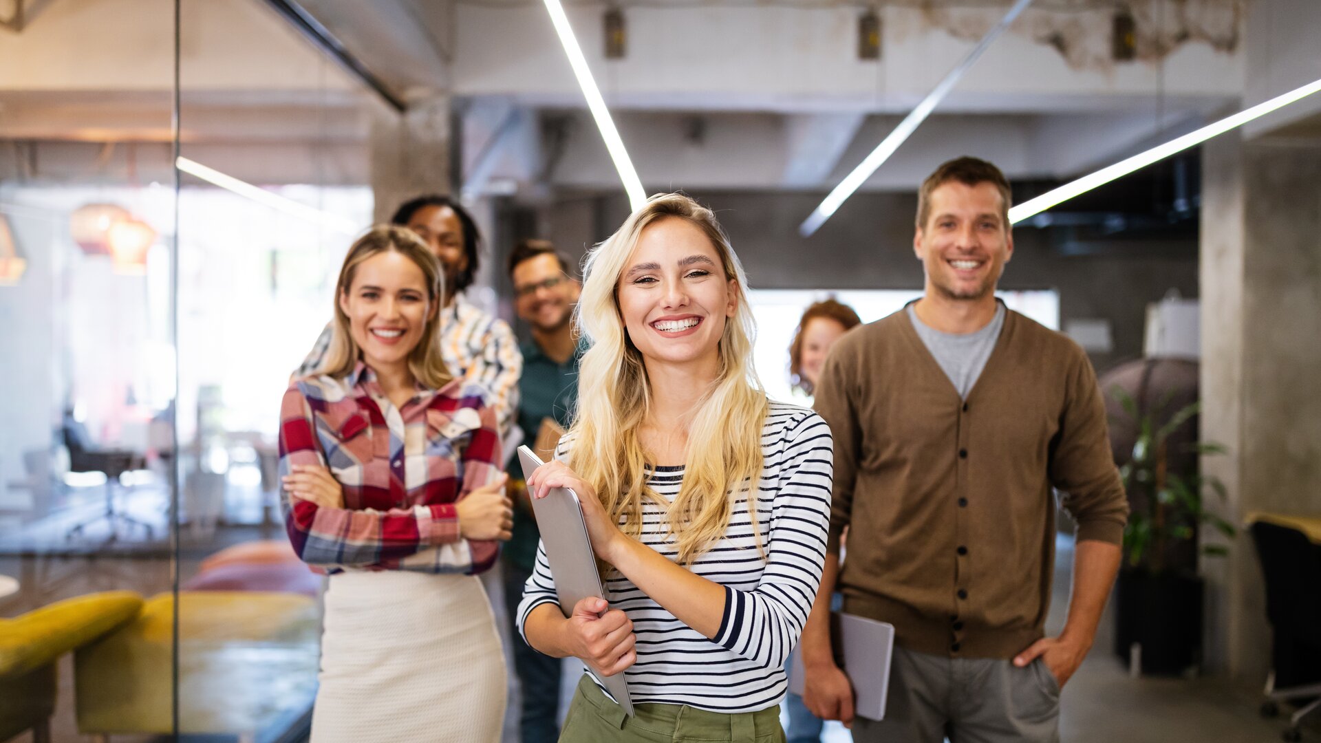Eine fröhliche Gruppe junger Menschen steht gemeinsam in einem modernen Büro und wirkt motiviert und gut gelaunt.