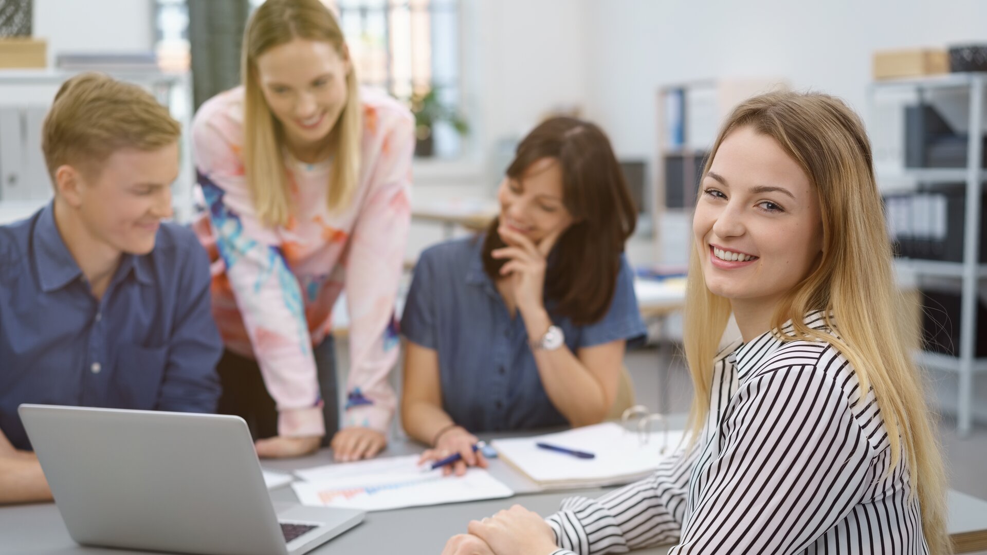 Eine junge Frau lächelt in die Kamera, während sie mit ihrem Team gemeinsam an einem Laptop arbeitet.