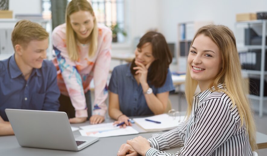 Eine junge Frau lächelt in die Kamera, während sie mit ihrem Team gemeinsam an einem Laptop arbeitet.