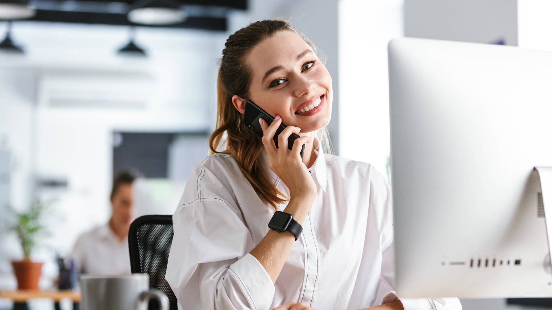 Eine lächelnde Frau telefoniert an ihrem Schreibtisch im Büro und arbeitet gleichzeitig am Computer.