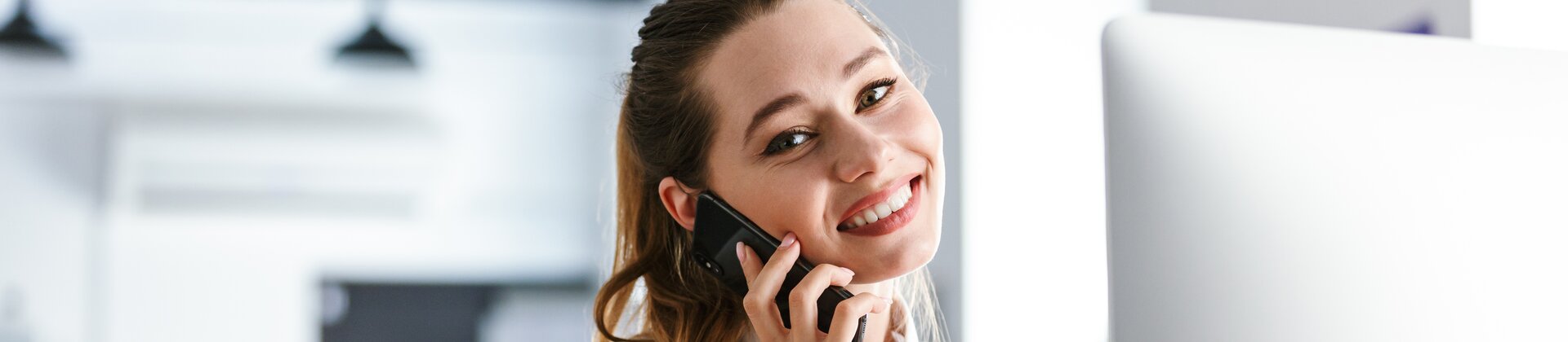 Eine lächelnde Frau telefoniert an ihrem Schreibtisch im Büro und arbeitet gleichzeitig am Computer.