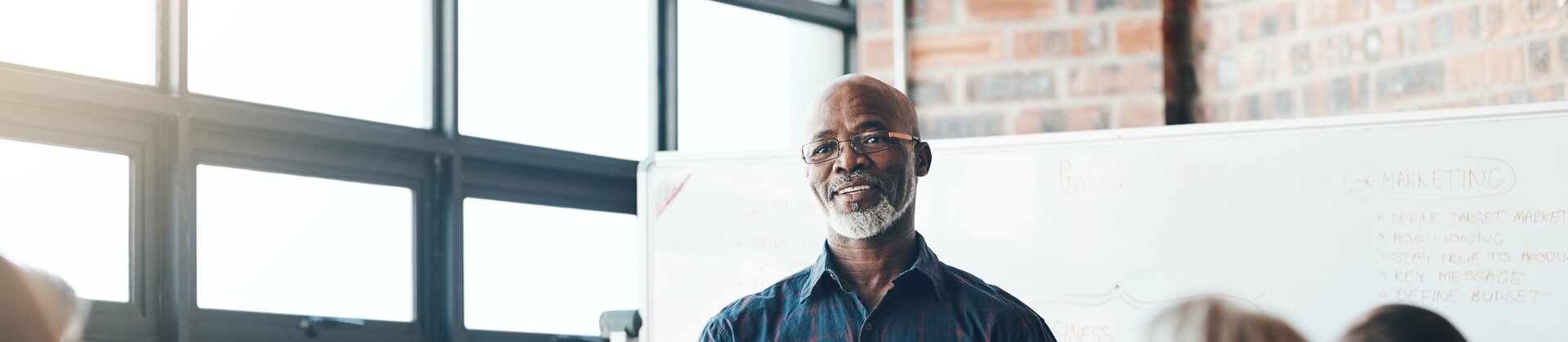 Ein Mann steht vor einem Whiteboard und erklärt etwas, während mehrere Personen aufmerksam am Tisch sitzen und ihm zuhören.