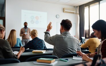 Eine Person hebt in einem Seminarraum die Hand, während der Vortragende vorne präsentiert und andere Teilnehmende mitschreiben.
