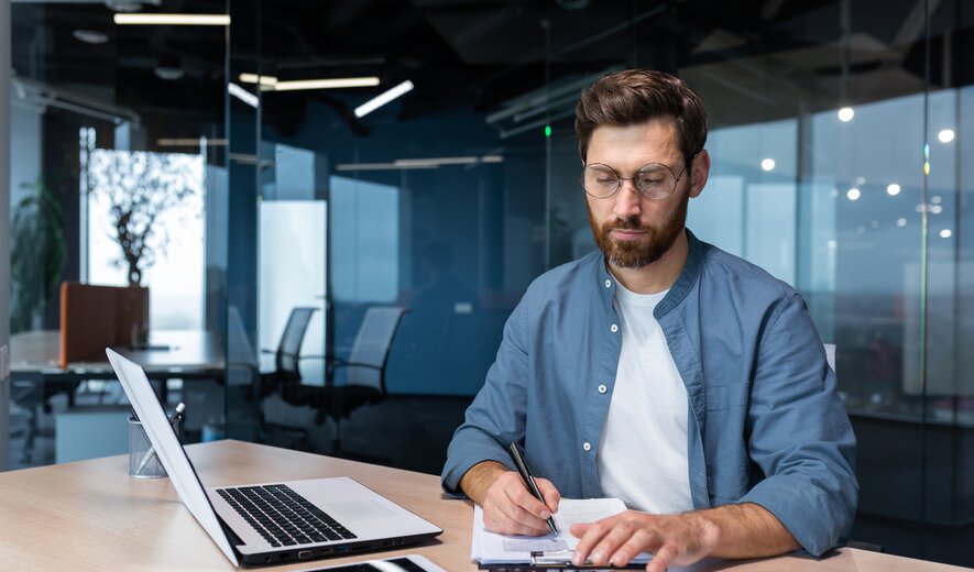 Ein Mann arbeitet konzentriert an seinem Schreibtisch, nutzt Laptop, Notizen und Taschenrechner für seine Aufgaben.