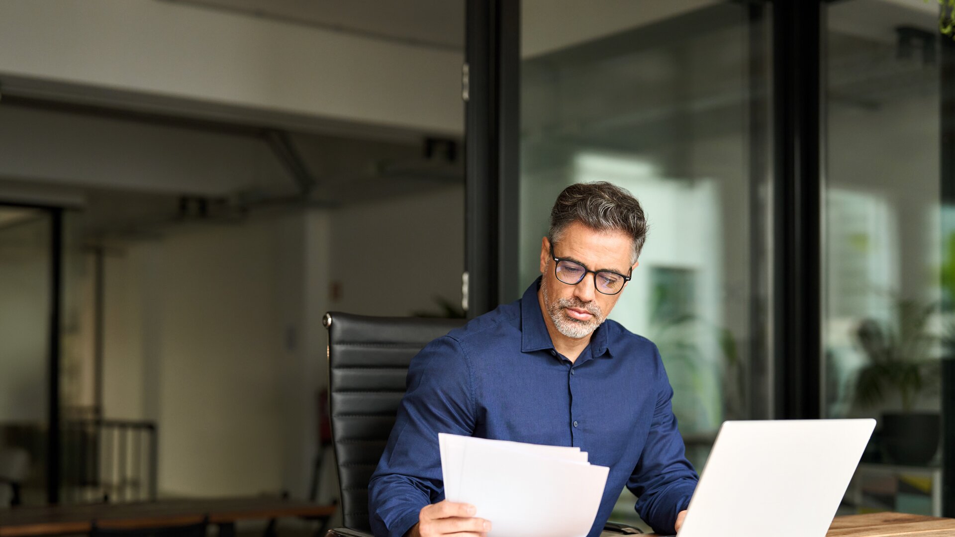 Ein Mann arbeitet konzentriert am Schreibtisch und prüft Unterlagen neben seinem Laptop.
