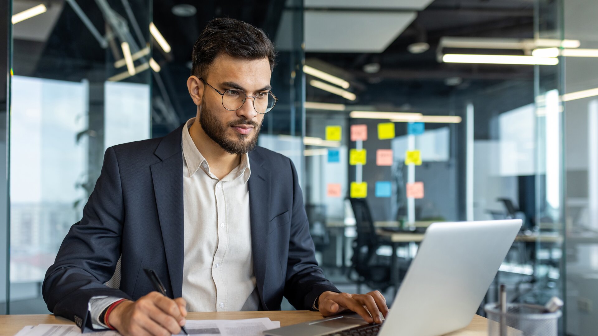 Ein Mann arbeitet konzentriert an seinem Laptop und macht sich dabei Notizen.