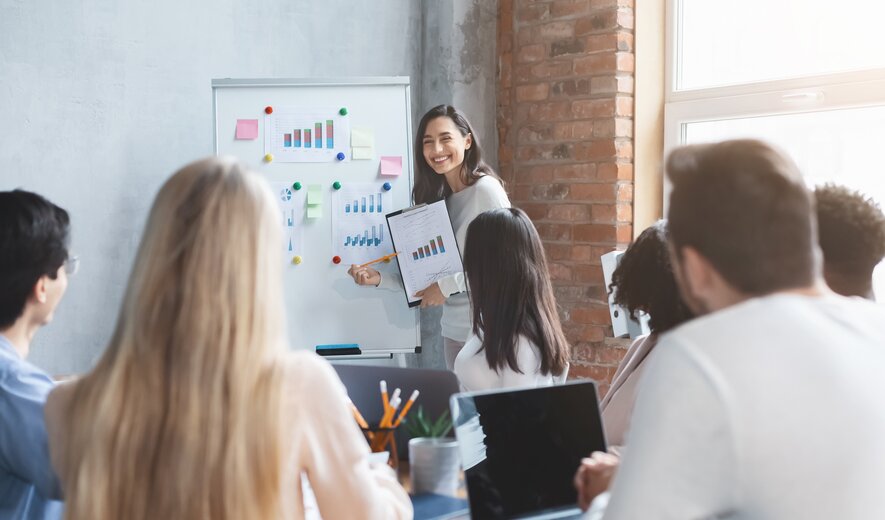 Eine Frau präsentiert lächelnd Grafiken vor einer Gruppe aufmerksam zuhörender Kolleginnen und Kollegen.